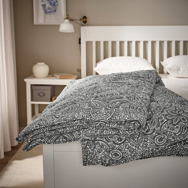 Un dormitorio minimalista con cama blanca, edredón gris, cabecero de madera y mesita de noche con lámpara, jarrón y libros.