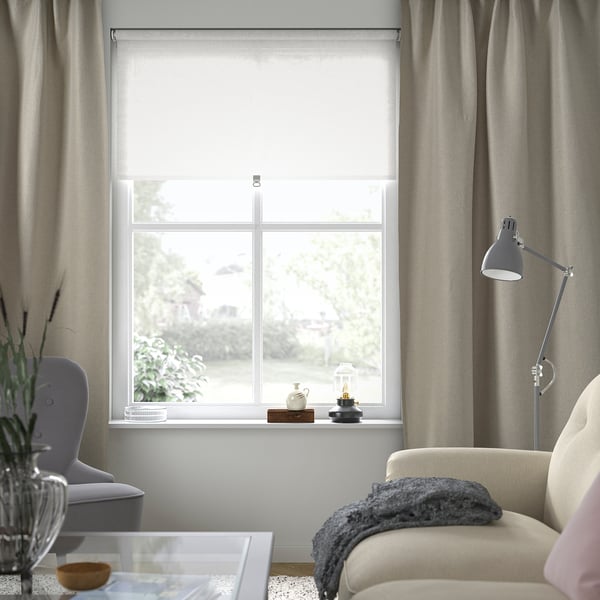 Acogedora sala de estar con cortinas grises, sofá beige, manta azul, lámpara de pie, silla gris, jarrón y mesa de centro de cristal.