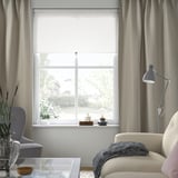 Acogedora sala de estar con cortinas grises, sofá beige, manta azul, lámpara de pie, silla gris, jarrón y mesa de centro de cristal.