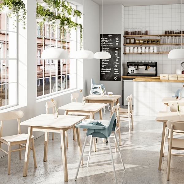 Cafetería moderna con mesas de madera, grandes ventanales, plantas colgantes y barra de café.