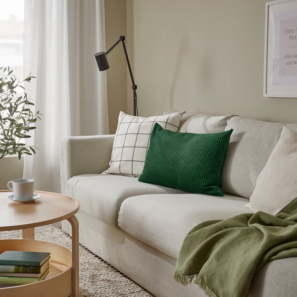 Acogedora sala de estar con un gran cojín blanco en un sofá claro, manta verde, mesa auxiliar con taza, libro y planta, y lámpara de pared.