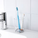 Dos cepillos de dientes en soporte transparente sobre la encimera del baño, junto a un grifo cromado. El soporte tiene pequeños pies de plástico para mayor estabilidad y protección.