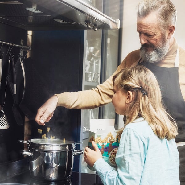 Un adulto y un niño cocinan pasta con forma de alce en la estufa, de una caja etiquetada como BÄSTISAR.
