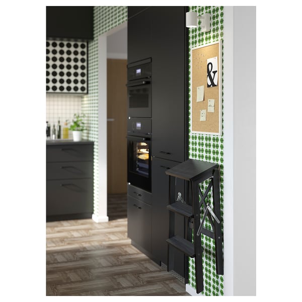 Cocina moderna con armarios negros, papel pintado verde estampado y una escalera de tijera plegable de madera de haya negra.