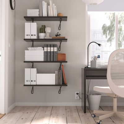 Moderno rincón de oficina con estanterías negras BERGSHULT que contienen libros, cajas y plantas. Un escritorio blanco, una silla y una gran ventana.