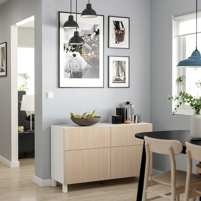 Comedor moderno con aparador de madera de color claro, encimera blanca, cafetera de acero inoxidable, luces colgantes. La pared tiene fotos.
