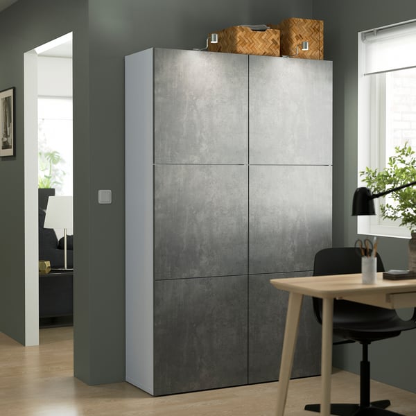 Estantería gris BESTÅ con puertas texturizadas, más alta que el escritorio y la silla, pared azul verdosa.