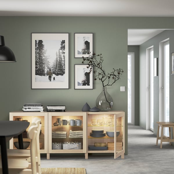 Comedor moderno con aparador de madera, puertas de vidrio, paredes de salvia, arte, luz suave.