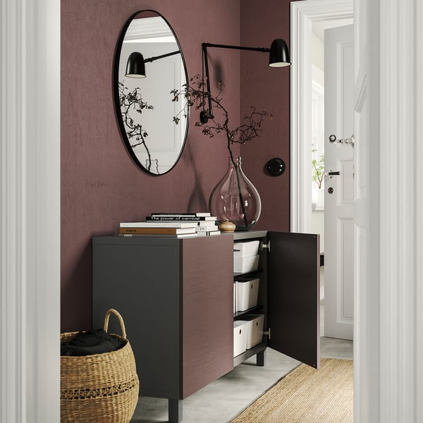 Armario BESTÅ de color marrón oscuro, con puertas abiertas que muestran organizadores blancos, contra una pared de color púrpura. Cubierto con libros, un jarrón y un gran espejo redondo.