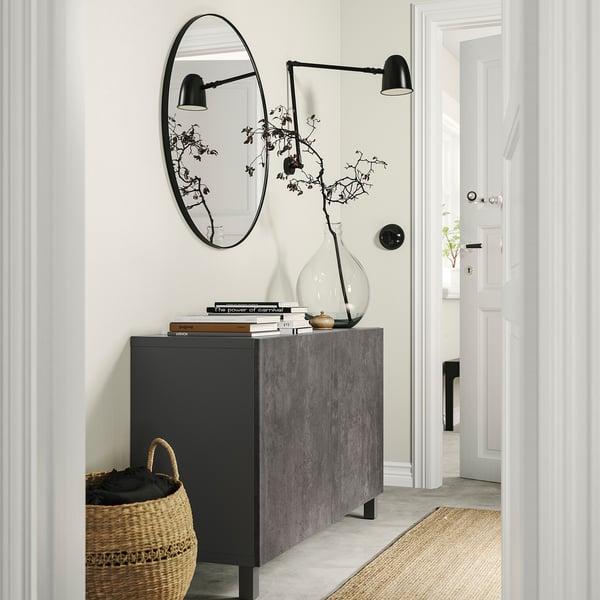 Entrada moderna con mueble de almacenamiento BESTÅ negro, espejo redondo, lámparas de pared, jarrón grande con ramas, libros y cesta tejida.