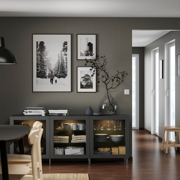 Habitación moderna con almacenamiento BESTÅ gris oscuro, lámpara colgante negra, fotos en blanco y negro, jarrones de vidrio, sillas de madera, pisos de madera y pasillo blanco.