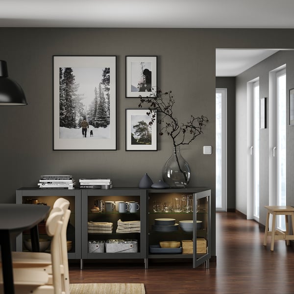 Comedor moderno con almacenamiento BESTÅ, puertas de vidrio, platos, jarrón con rama desnuda, debajo de fotos en blanco y negro.