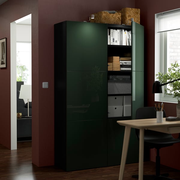 Armario BESTÅ verde oscuro con estantes ajustables, almacenamiento de libros y cajas. Puertas de cierre suave, colocadas junto a una mesa y una silla de madera.