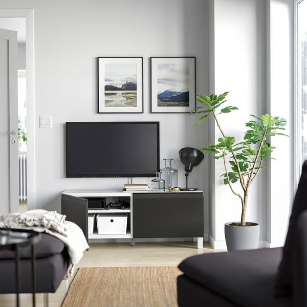 Sala de estar moderna con un mueble de TV BESTÅ negro, TV de pantalla plana, dos cuadros de naturaleza enmarcados, una planta verde alta y paredes grises.