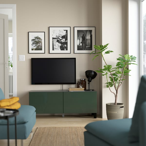 Sala de estar moderna con mueble de TV verde BESTÅ, estantes ajustables y fotos de pared. Una planta alta y verde complementa un sillón verde azulado.