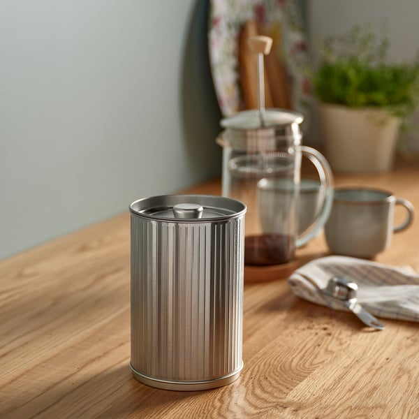 Bote moderno, metálico y cilíndrico sobre una mesa de madera. Más pequeña que la cafetera y las tazas, tiene un diseño acanalado, acabado plateado.