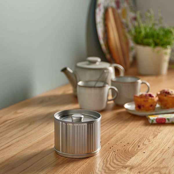 Contenedor metálico compacto BETTHAJ, plateado, se coloca en el centro sobre una mesa de madera, más pequeña que tazas. Cerca de tetera y magdalenas.