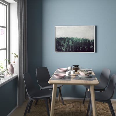 Un comedor moderno con una mesa de madera y sillas negras. La pared azul tiene un póster enmarcado del bosque. Hay una ventana con un jarrón de flores.