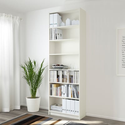 Librería blanca BILLY con libros y cajas, cerca de ventana, planta verde, alfombra a rayas.