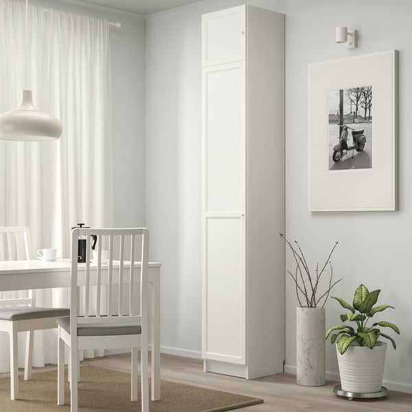 Área de comedor minimalista con estantería blanca BILLY, planta en maceta, foto de scooter enmarcada y sillas blancas alrededor de una mesa.