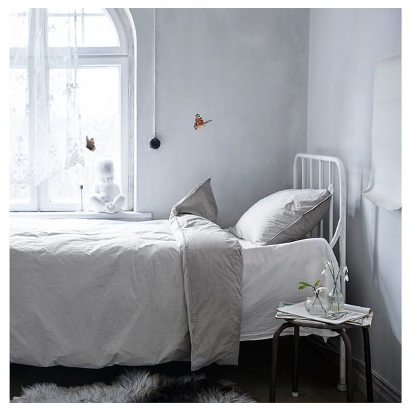 Dormitorio minimalista con cama BLÅVINDA, edredón de rayas grises, almohadas blancas, estructura metálica. Mesa auxiliar con vegetación cerca de la ventana.