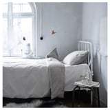 Dormitorio minimalista con cama BLÅVINDA, edredón de rayas grises, almohadas blancas, estructura metálica. Mesa auxiliar con vegetación cerca de la ventana.