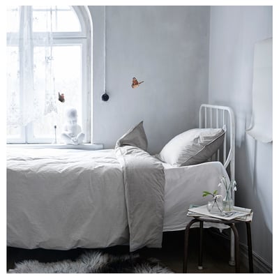 Dormitorio minimalista con cama BLÅVINDA, edredón de rayas grises, almohadas blancas, estructura metálica. Mesa auxiliar con vegetación cerca de la ventana.