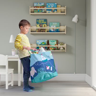 Niño con camisa a rayas sosteniendo una bolsa de almacenamiento con temática oceánica. Detrás de él hay estanterías llenas de coloridos libros infantiles.