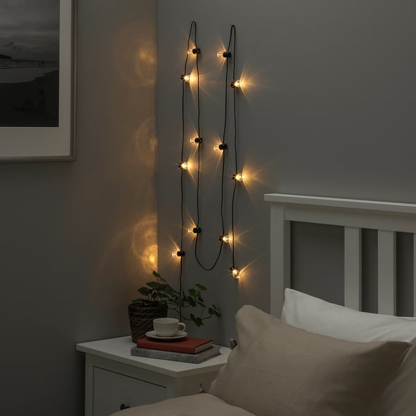 Acogedor dormitorio con guirnaldas de luces, cama, mesita de noche con taza, libros y plantas.