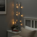 Acogedor dormitorio con guirnaldas de luces, cama, mesita de noche con taza, libros y plantas.