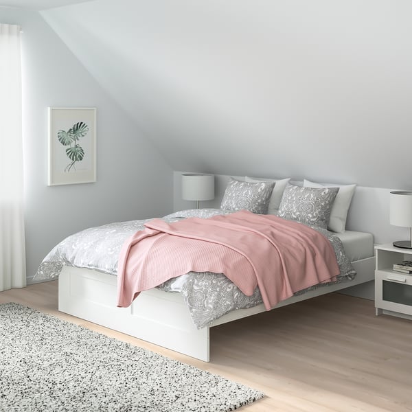 Cama minimalista BRIMNES blanca, manta rosa claro, almohadas grises, junto a la ventana, mesitas de noche pequeñas. Se adapta a espacios compactos.