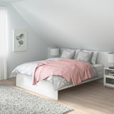 Cama minimalista BRIMNES blanca, manta rosa claro, almohadas grises, junto a la ventana, mesitas de noche pequeñas. Se adapta a espacios compactos.