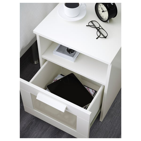 Mesita de noche blanca con cajón abierto que muestra los artículos almacenados: tableta, libro y cable. La parte superior sostiene una taza de café, un platillo, vasos y un despertador.