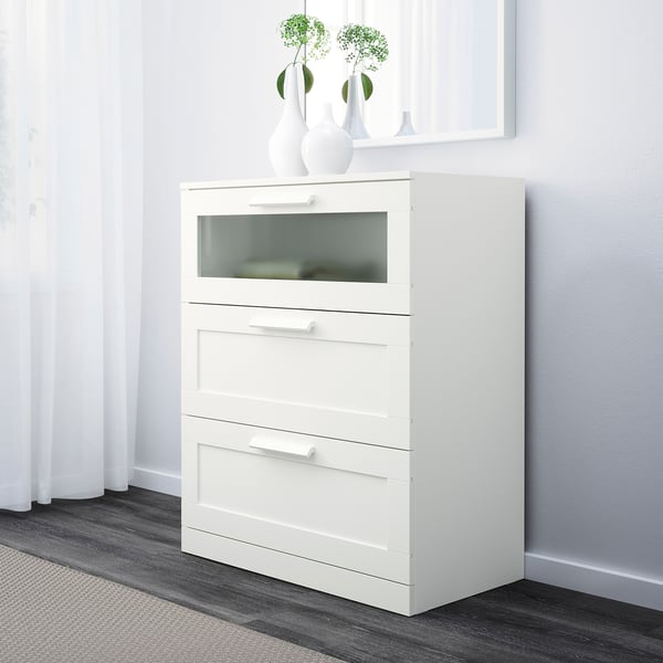 Armario blanco con cajón superior de cristal, dos cajones normales y jarrones decorativos en la parte superior, colocados contra una pared ligera.