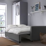 Dormitorio moderno y minimalista con sofá cama gris BRISSUND, ropa de cama blanca, prenda colgante, estantería de pared con decoración y luz natural por la ventana.