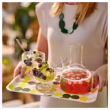 Persona sosteniendo la bandeja con puntos amarillos. La bandeja tiene postres de frutas y bebida con hielo. El borde en ángulo mantiene los artículos seguros.