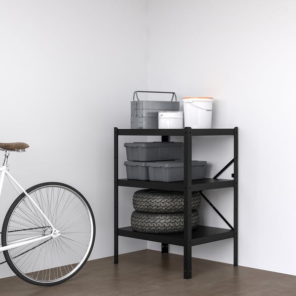 Estante de almacenamiento de metal negro BRA. Sostiene neumáticos, contenedores y cubos. Bicicleta inclinada cerca, se adapta a espacios reducidos.