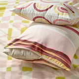 Sobre una cama descansan dos fundas de almohada de diseño abstracto en suaves tonos rosa y verde, una de ellas con cremallera para facilitar su extracción, fabricada con poliéster reciclado.