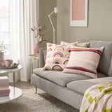 Acogedora sala de estar con sofá gris, cojines BRUNTÅG rosas y beige, mesa auxiliar con planta y taza, lámpara cálida y manta con detalles verdes.