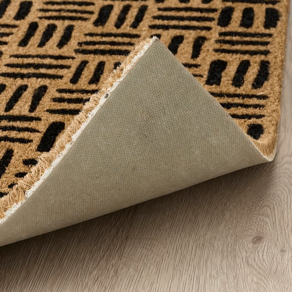 El felpudo de fibra de coco natural con rayas negras descansa sobre el suelo de madera, mostrando su respaldo texturizado diseñado para mantenerlo firmemente en su lugar.