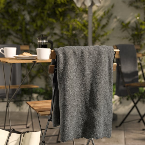 Manta de vellón DAGGKÅPOR gris oscuro sobre la silla, más grande que tazas y platos sobre una mesa de madera cerca de plantas verdes.
