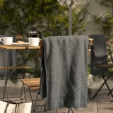 Manta de vellón DAGGKÅPOR gris oscuro sobre la silla, más grande que tazas y platos sobre una mesa de madera cerca de plantas verdes.