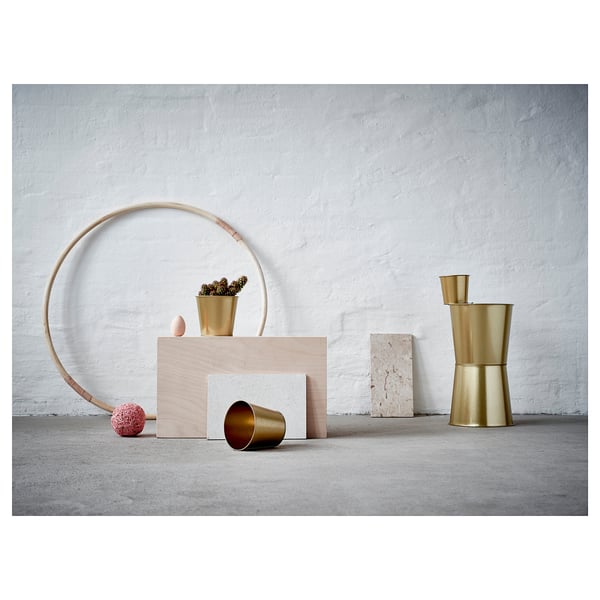 Escena minimalista moderna con macetas de metal de color latón, una decoración circular de madera y bloques geométricos apilados contra una pared con textura blanca.