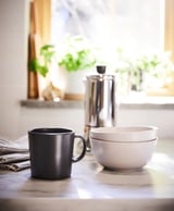 Una configuración de cocina moderna con taza y tazón de gres blanco y negro, una prensa francesa y plantas verdes sobre una encimera de mármol.