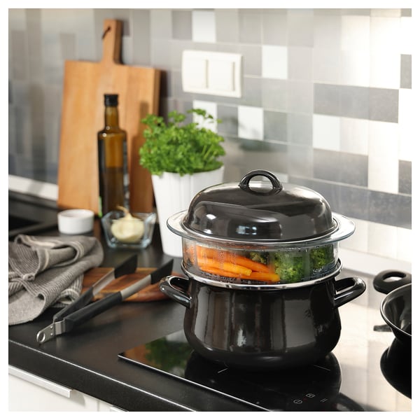 Cazuela negra con tapa de vidrio, verduras al vapor. Cocina moderna con utensilios y hierbas.