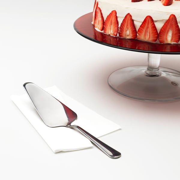 Un rebanador de pastel brillante en una servilleta, listo para cortar un pastel cubierto de fresa en un soporte de vidrio.