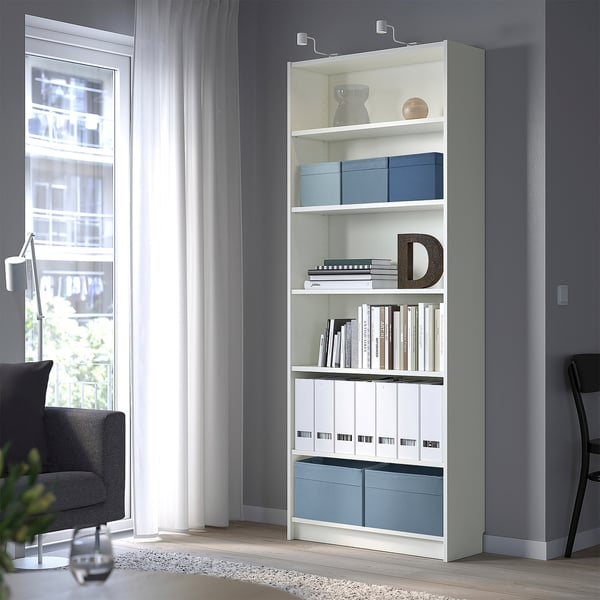 Estantería blanca con cajas, jarrones y libros azules. La habitación tiene silla, ventana, lámpara.