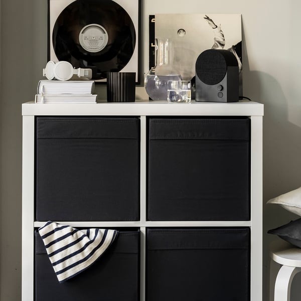 Estantería blanca con cajas de almacenamiento de tela negra DRÖNA, disco de vinilo, libros, altavoz y gorro a rayas.