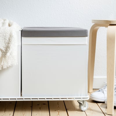 Tapa de caja de tela gris sobre suelo de madera con silla y perchero.