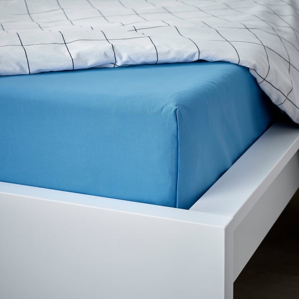 Una cama con una sábana plana DVALA de algodón azul, ligeramente doblada sobre un marco blanco, que promete comodidad y transpirabilidad.